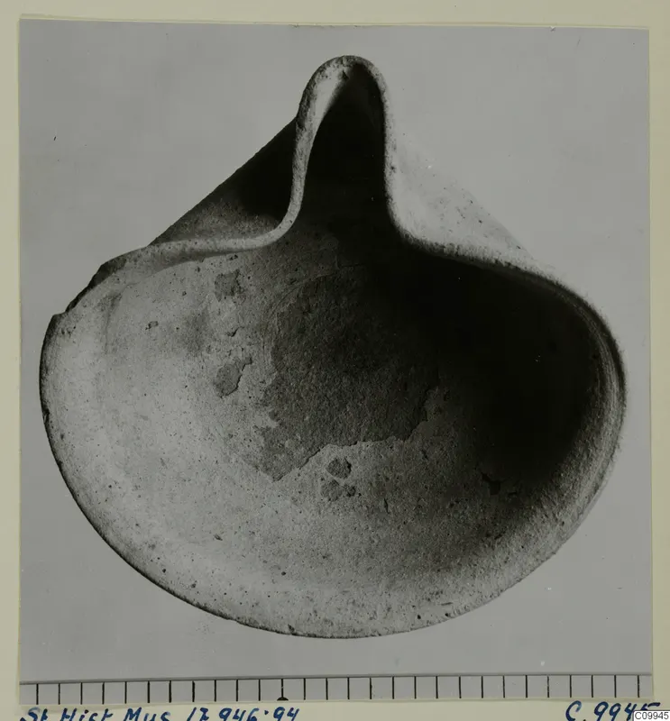 Terracotta lamp with a shallow rounded body and a pinched-up nozzle, shown against a grey background beside a scale bar.