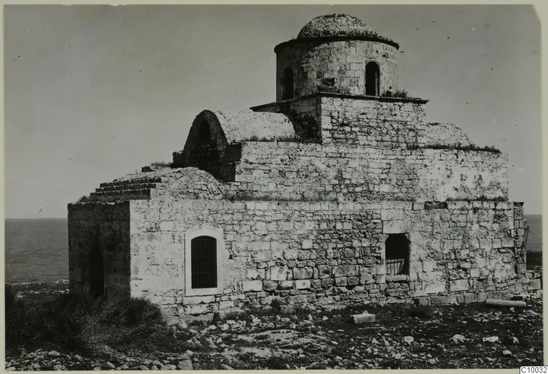 Monastery building of stone with a dome on a drum and arched windows, with the sea behind.