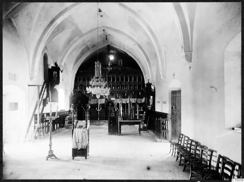 Interior of a monastery church looking toward a dark wooden screen with icons, with chairs along the right wall and a wooden door.