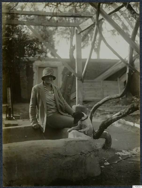 A smiling woman in a cloche hat sits on a large stone trough beside a dog, under a timber frame.