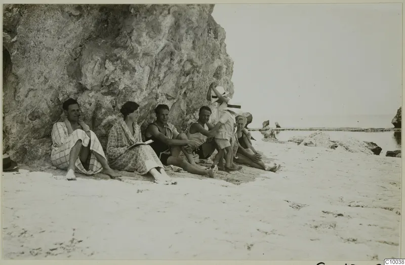 Group of seven people sitting against a rock face on a sandy beach by the sea.