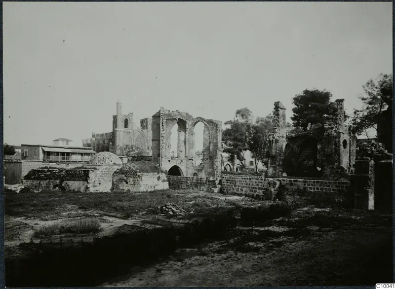Ruined stone buildings with tall arched openings and a slender tower, Famagusta, seen across grass and low walls.