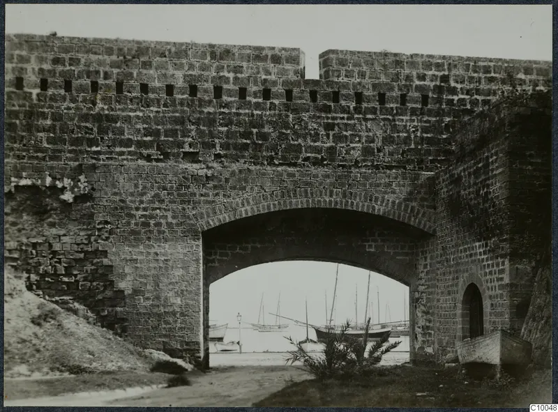 Arched gateway through a masonry fortification wall, with sailing boats moored in the harbour visible through the arch.