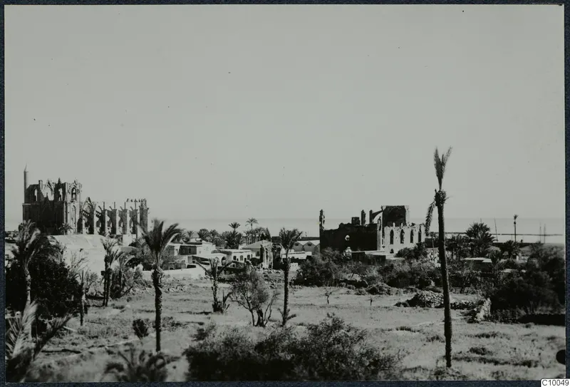 Ruined church walls with pointed window openings among palm trees and low houses at Famagusta, with the sea beyond.