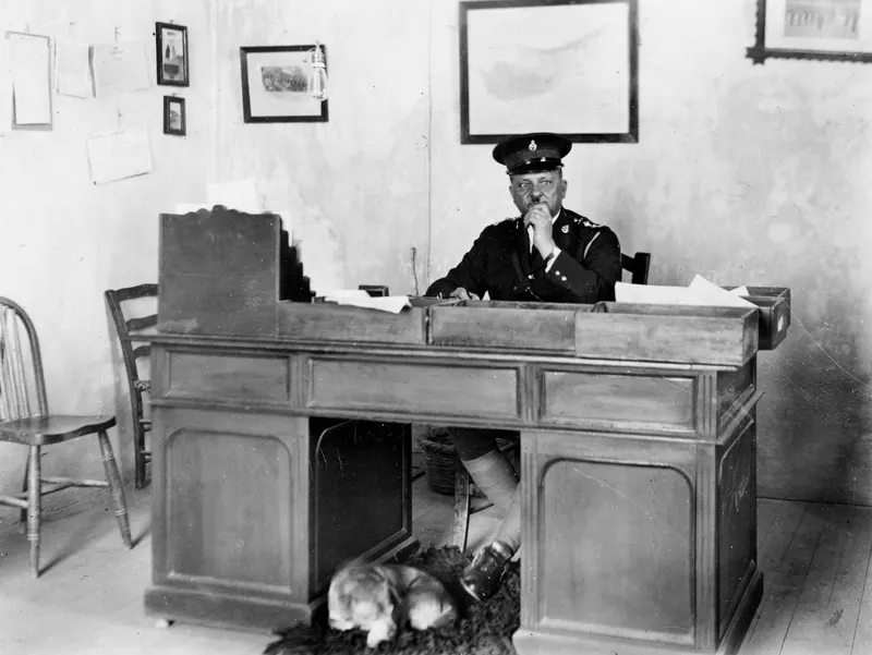 Man in a dark uniform and peaked cap seated at a wooden desk, with a dog lying on a rug beneath it, Famagusta.