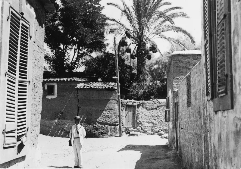 Ernst Kjellberg standing in a sunlit village street between mud-walled houses, with a palm tree behind.