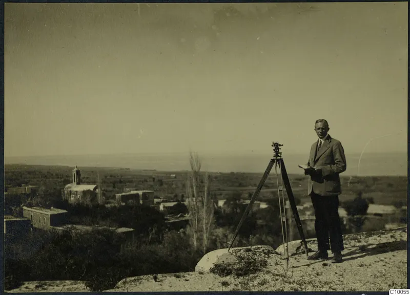 Man in a suit beside a tripod-mounted instrument on a rise, with a church, village and the sea beyond.