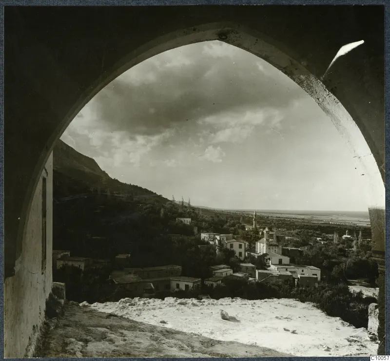 View through a stone arch over a hillside village of pale houses and trees, with a steep mountain slope on the left.