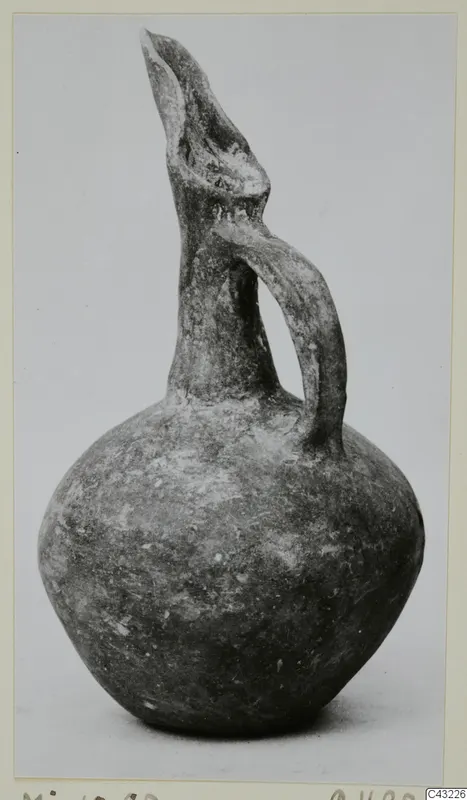 Dark grey-brown juglet with a tall trefoil-shaped spout and a single loop handle, photographed on a plain background.