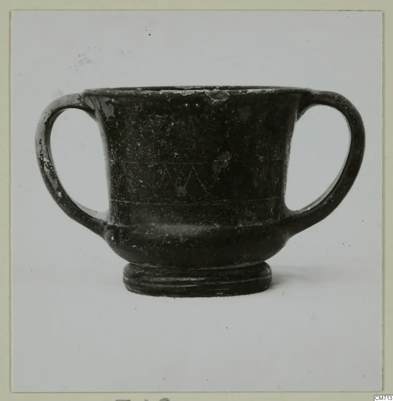 Two-handled dark glazed vessel with incised horizontal lines and a zigzag band on its body, seen against a plain background.