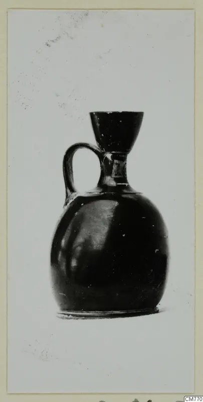 Small black-glazed jug with a single loop handle and a funnel-shaped mouth, photographed against a pale background