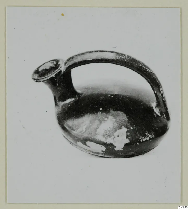 Black-glazed pottery jug with a spout and a strap handle, photographed on a plain background