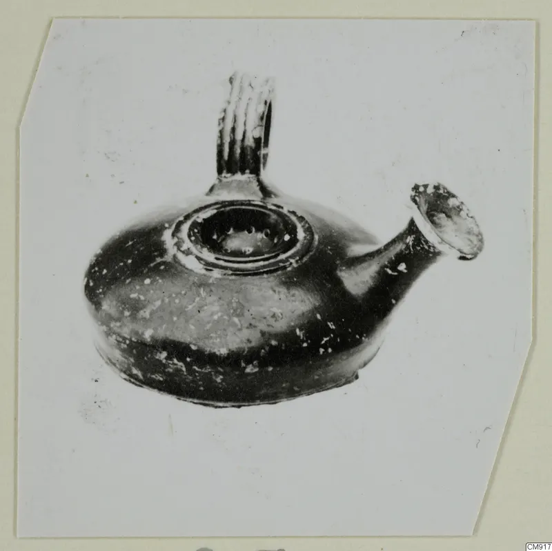 Black-glazed pottery oil lamp with a ribbed handle and a spout, photographed on a card mount