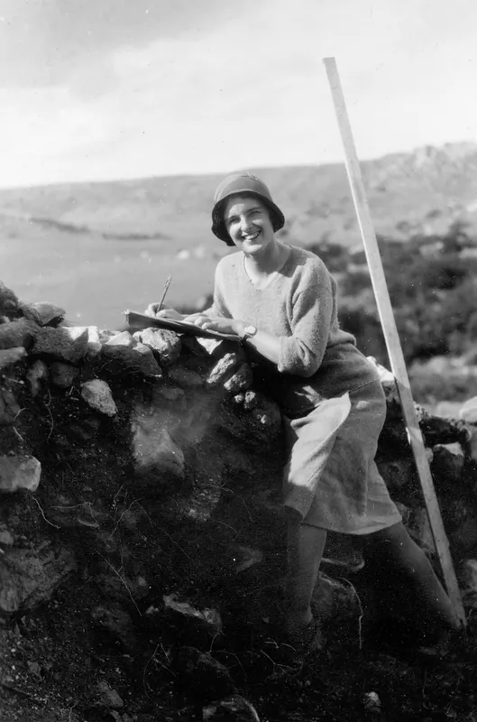 Ebba Elgenstierna seated on a stone wall, writing on a clipboard, with a wooden measuring rod leaning beside her at Nitovikla