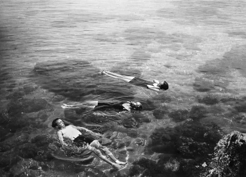 Three people floating on their backs in clear sea water beside a rocky outcrop.