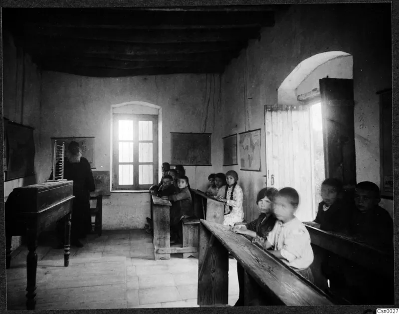 Classroom with a bearded teacher in a long dark robe standing by a desk, and children seated on wooden benches.
