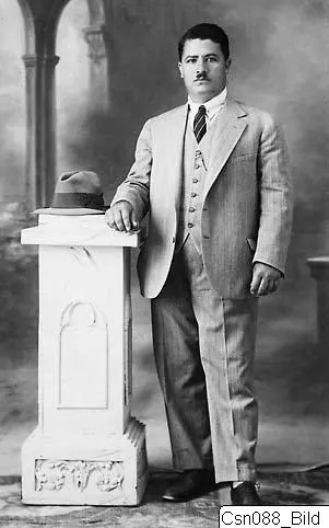 Studio portrait of Toulis in a light suit, standing beside a white pedestal with a hat on top