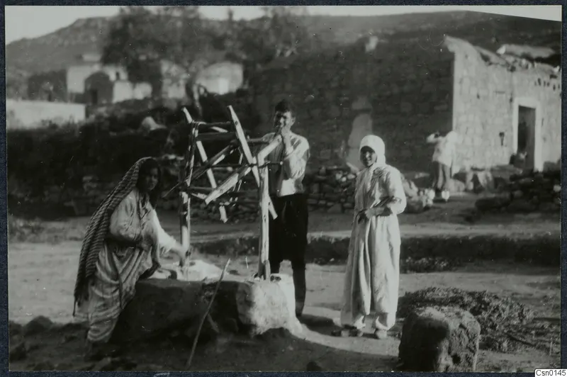 Three people beside a stone well with a wooden frame in a village street, with a stone building behind them