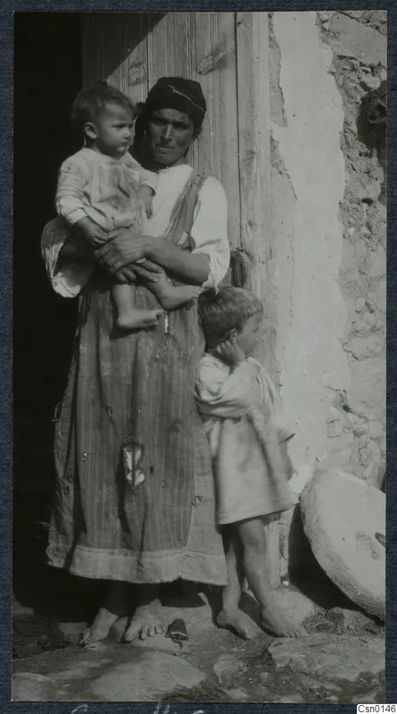 Woman in a headscarf holding a toddler in a doorway, with a small child standing beside her against a stone wall