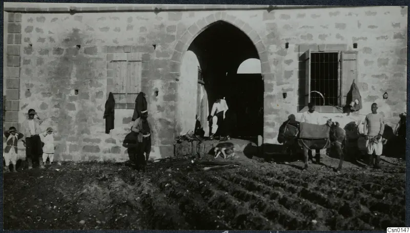Men, children and a saddled donkey before a stone building with a pointed-arch doorway, beside a ploughed field