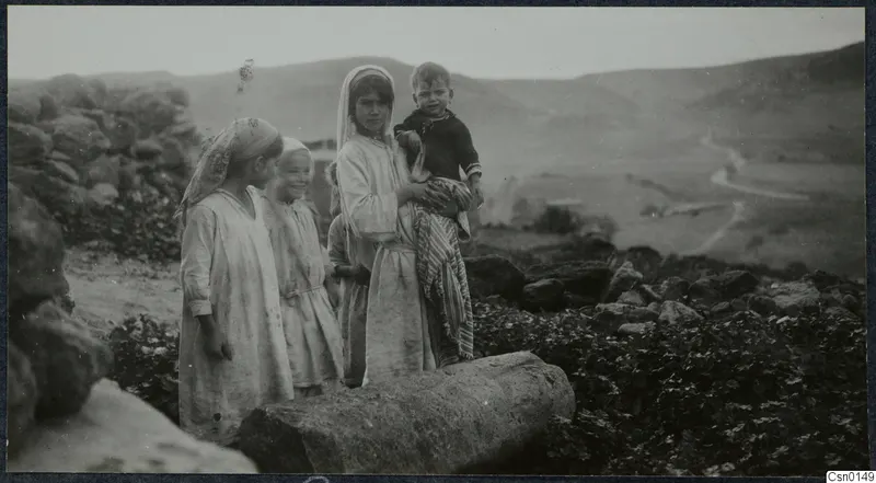 Children standing by a large cut stone block in a rocky field, with hills and a winding road behind
