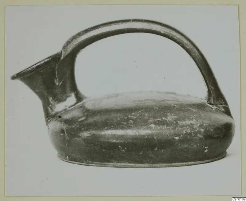 Dark-surfaced pottery jug with a high strap handle and a spout, photographed from the side on a plain background