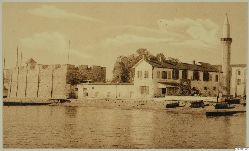 Waterfront with a stone fortification, a two-storey building and a minaret, with small boats drawn up on the shore