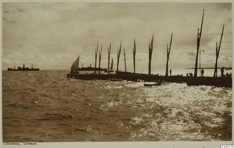 Long jetty at Limassol lined with bare masts, a sailing boat and a steamship beyond, and people standing on the pier over choppy sea