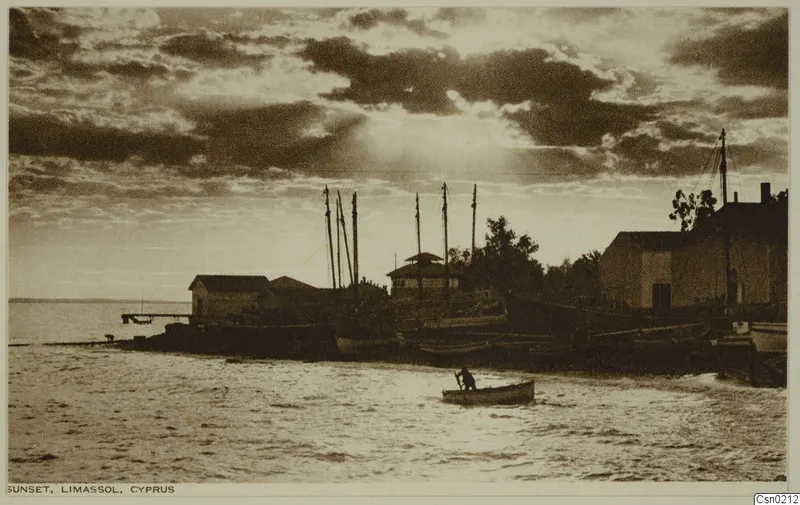 Sunset over the shore at Limassol, with boats with masts drawn up by white buildings and one person rowing a small boat.