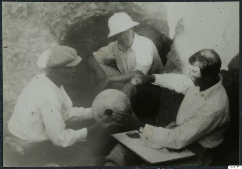 Three men handling a rounded object beside a dark rock-cut opening, one seated at a small board