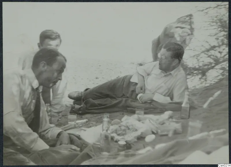 Three men resting on sand beside bottles, glasses and food spread on a cloth.