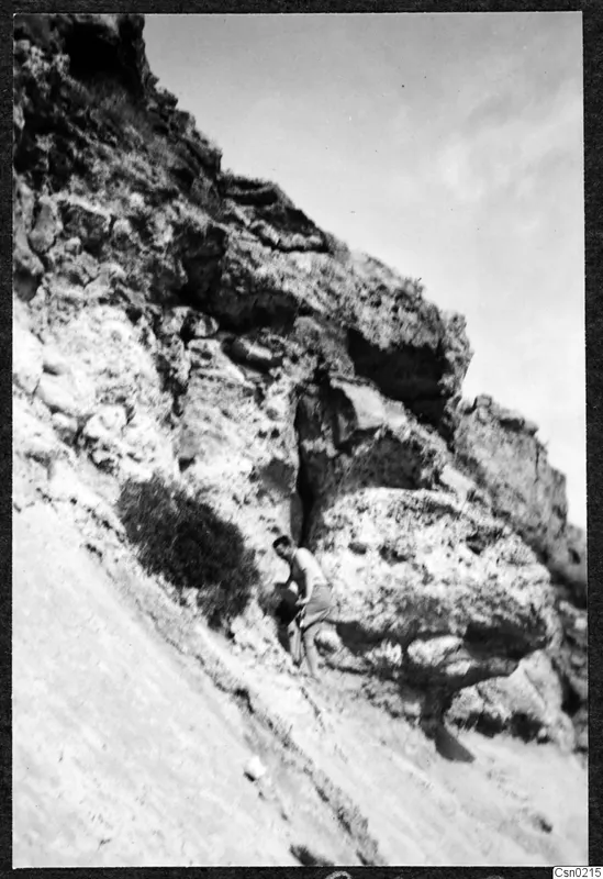 A man climbs a steep slope of weathered rock outcrops, with loose stones and scrub below.