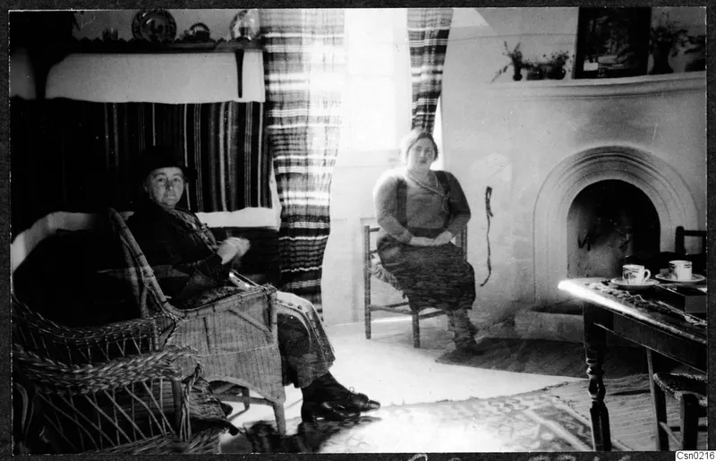 Two women seated in a room with a fireplace, a wicker armchair and a small table with cups and saucers
