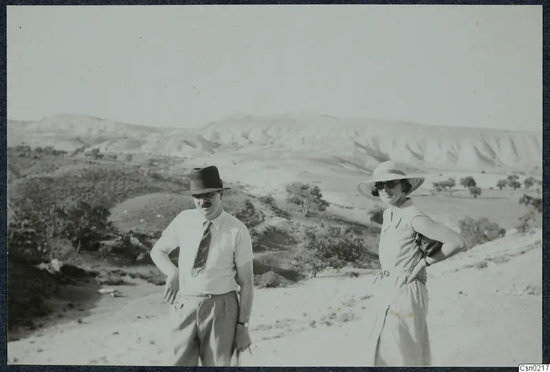 A man and a woman stand on a dusty track in front of pale hills, scrubby bushes and small trees.