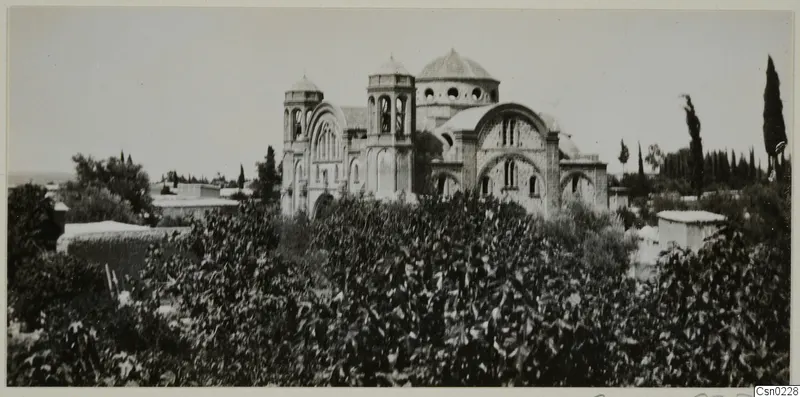Domed church with bell towers and arched windows, seen above dense shrubs, with cypress trees at the right