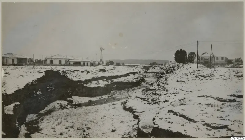Deep eroded gully in the foreground, with white buildings, a windmill, trees and low hills beyond at Nicosia.