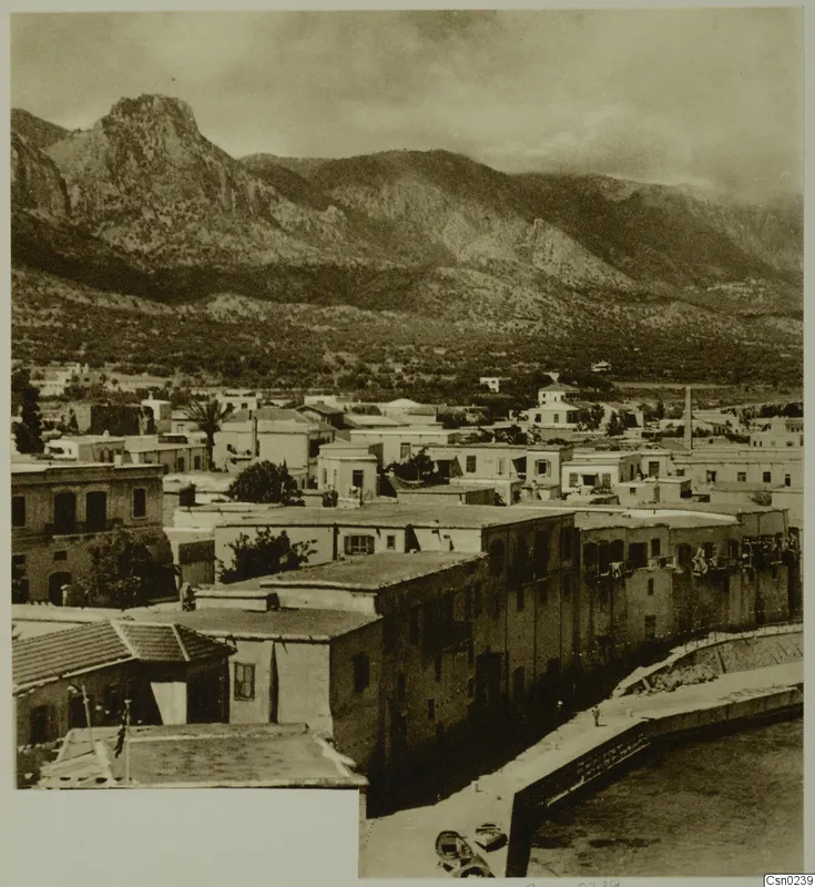 Flat-roofed houses of a town below steep mountains, with a harbour quay and small boats at the lower right