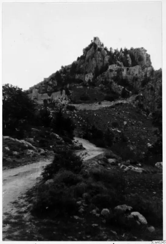 Dirt road through bushes below a rocky mountain with ruined stone walls on its slopes and summit, Kyrenia Mountains