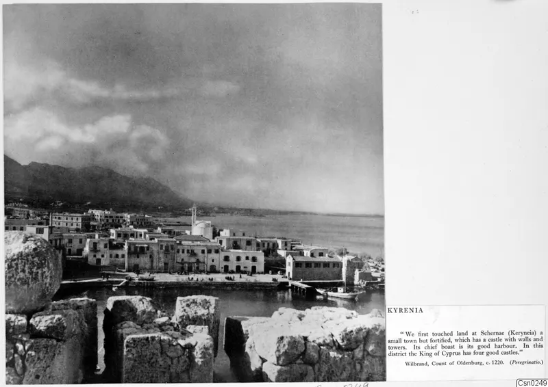Kyrenia harbour and waterfront seen over ruined stone blocks, with a moored boat and mountains behind the town.
