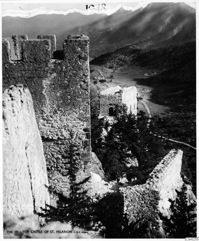 Ruined hilltop castle walls and a small stone building above a valley, with mountains behind