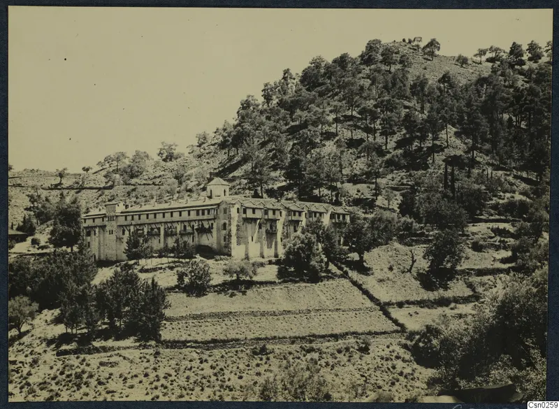 Long stone building with a small tower on a wooded hillside above terraced fields.
