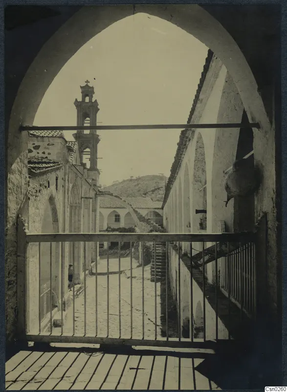Courtyard of buildings seen through a pointed arch, with a bell tower, arcaded walls and a stone staircase