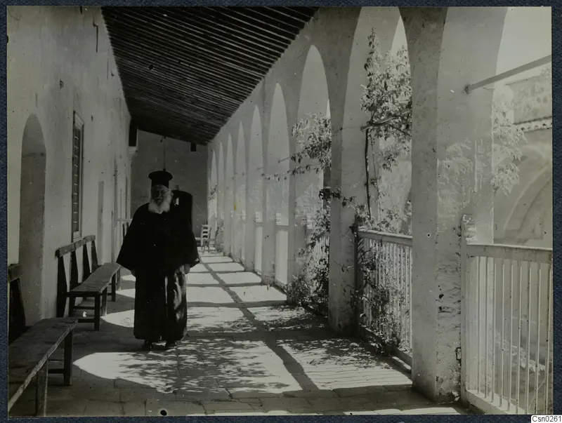 Bearded man in a long dark robe and tall black hat standing in a covered arcade with arches and a tiled floor.
