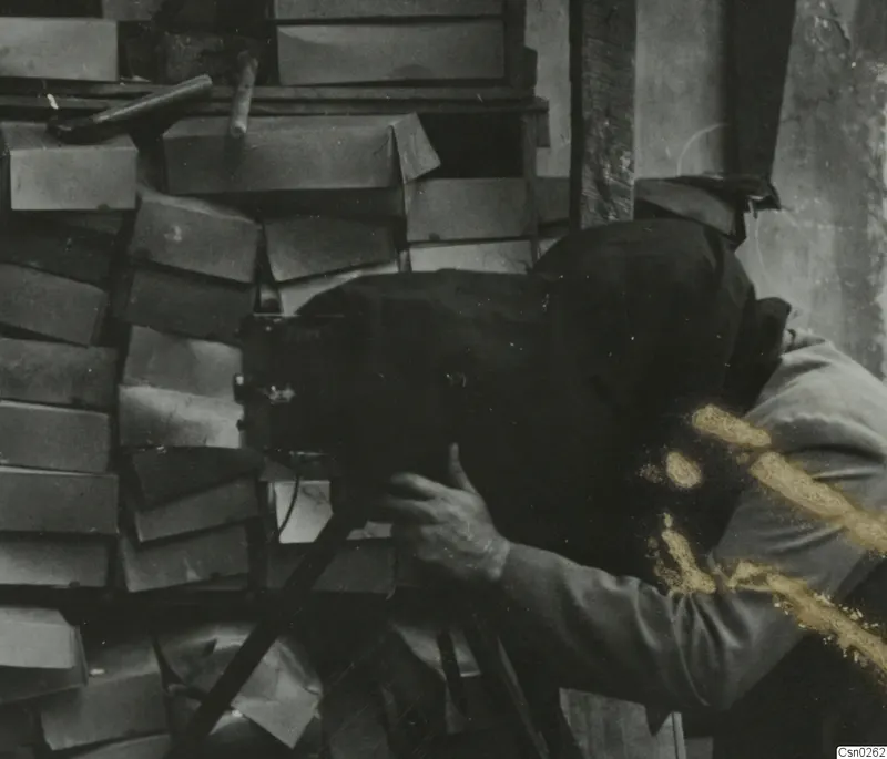Photographer with head under a dark cloth behind a large camera, with stacked wooden boxes behind.