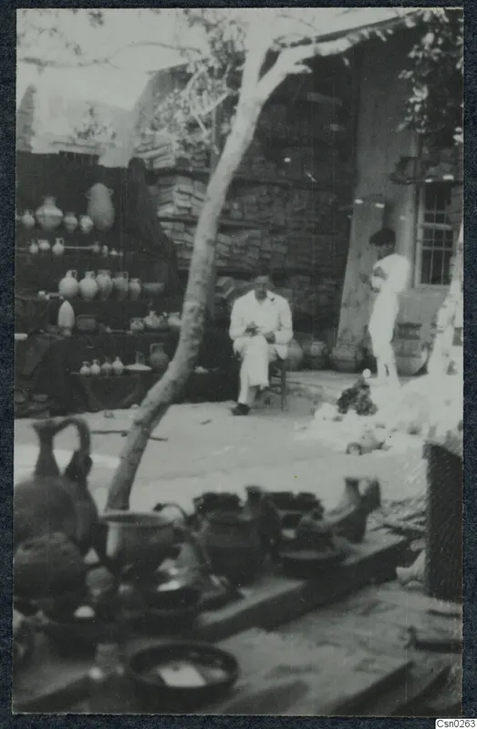 Seated man in a light suit beside shelves of pottery jugs and jars in a courtyard, with a standing figure to the right