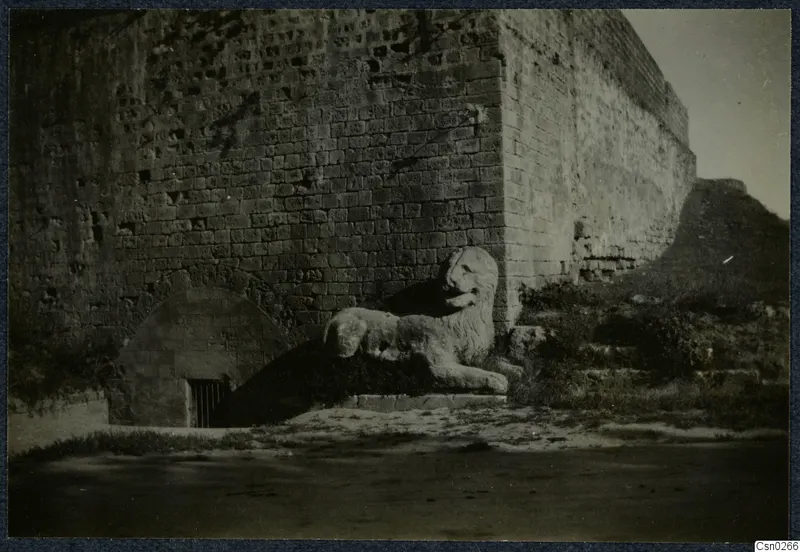 Stone lion sculpture lying beside a large masonry wall and an arched opening with a barred grate