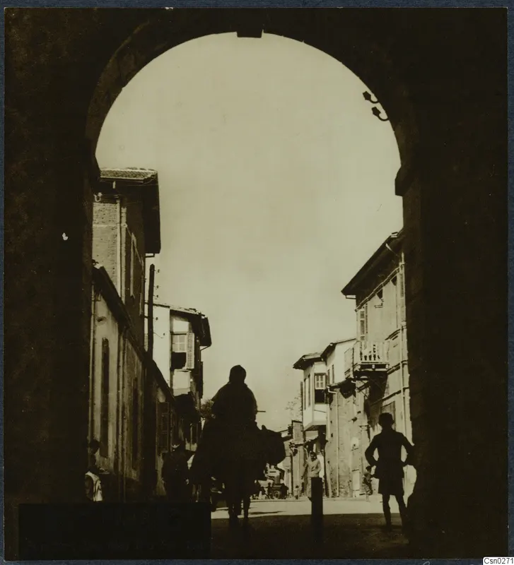 Rider silhouetted under a stone archway, looking down a street of tall buildings with balconies and shutters