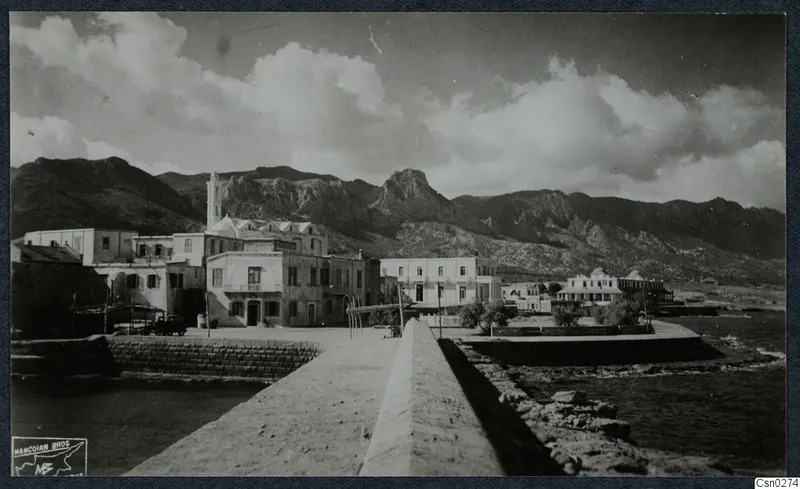 Seafront with a stone quay wall running towards plastered buildings, with rocky mountains and the sea behind and beside them