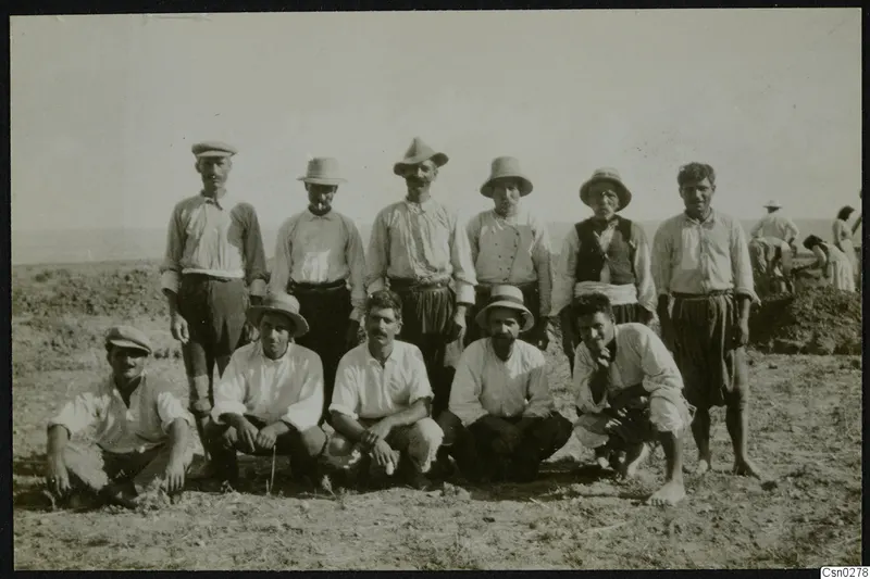 Group of men in hats and caps posing in a dry field, with other workers bending over earth in the background.