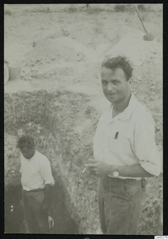 Two men at the edge of an excavation trench, one smiling at the camera, with spoil heaps behind them.